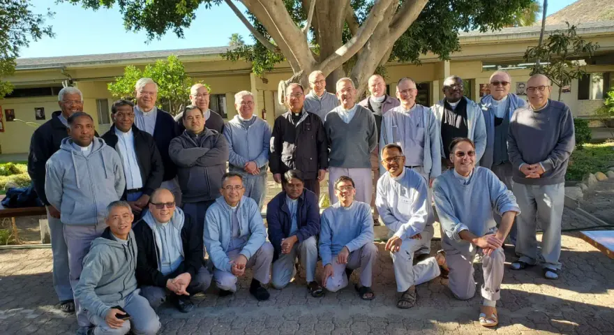 A group of missionaries of charity fathers pose for a photo outdoors, standing and kneeling under a large tree.