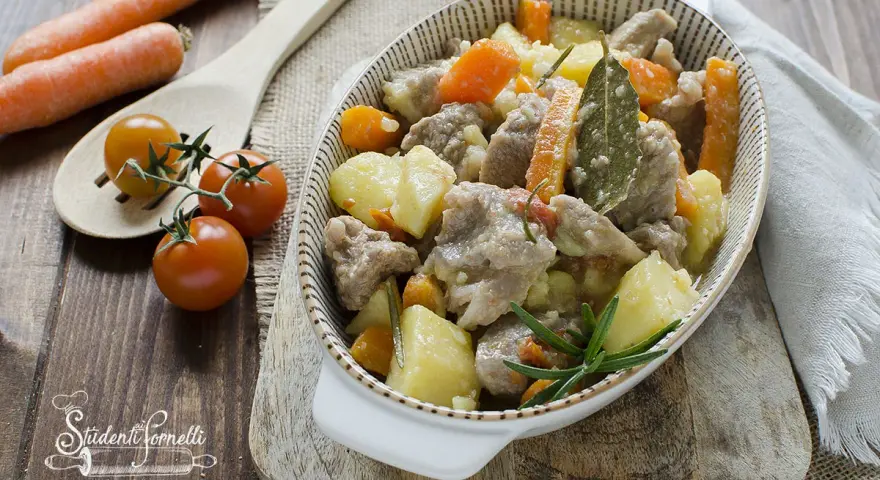 Un invitante spezzatino in bianco con patate e carote, guarnito con rosmarino e alloro. Accanto, pomodorini e carote fresche.