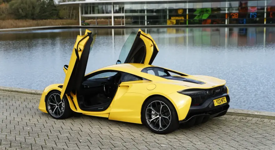 Żółty McLaren Artura z otwartymi drzwiami, stojący nad jeziorem przy nowoczesnym budynku.