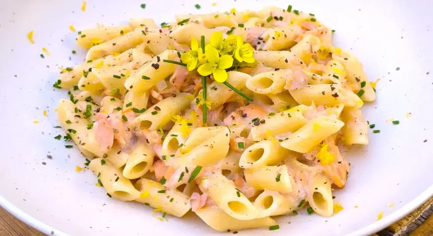 Pasta penne cremosa con trozos de salmón rosado, ralladura de limón y flores amarillas.