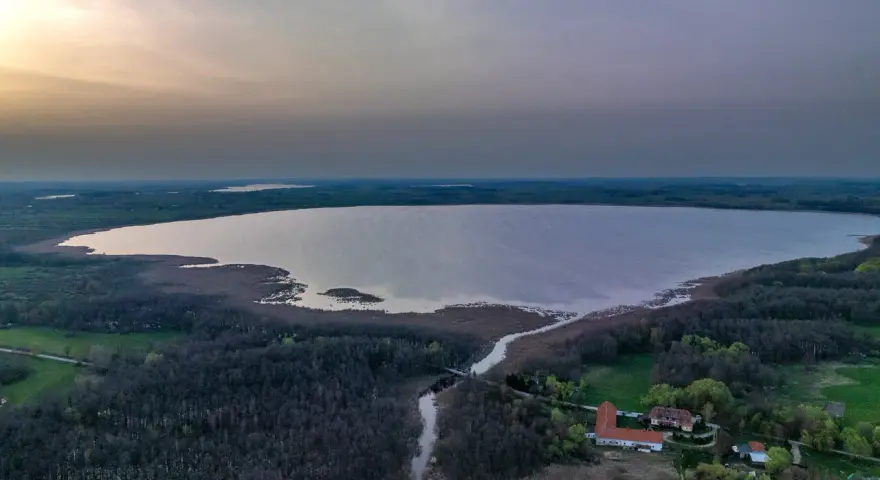 Rozległe jezioro Tuchlin otoczone lasem, z widocznym budynkiem i drogą. Zachód słońca maluje niebo.