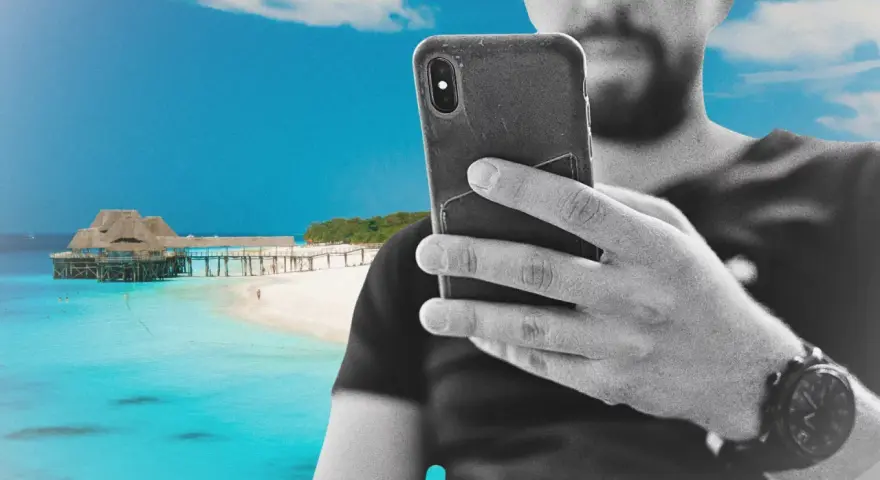 Mężczyzna z telefonem na plaży na Zanzibarze, sprawdzający zanzibar internet. W tle turkusowa woda i drewniany pomost.