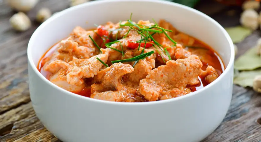 Pyszne wieprzowina w sosie curry, udekorowana szczypiorkiem i papryczką chili, podana w białej misce na drewnianym stole.