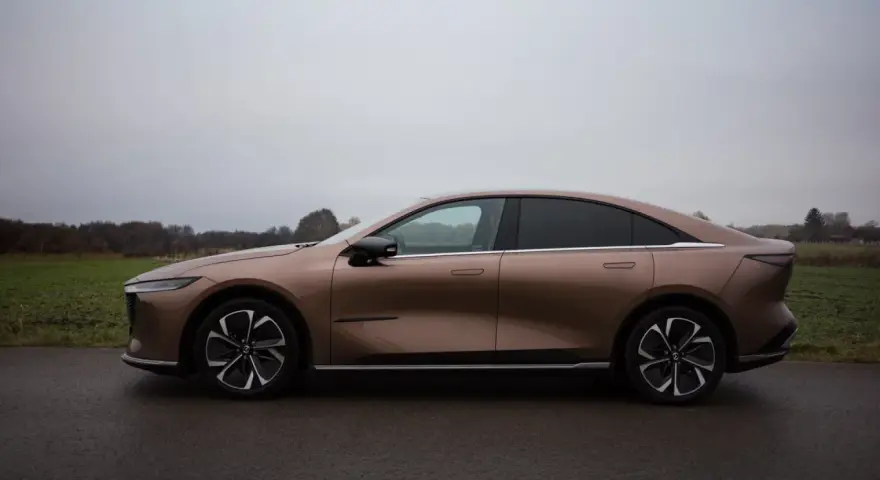 Brązowy samochód elektryczny Mazda6e widziany z boku na tle pochmurnego nieba i krajobrazu.