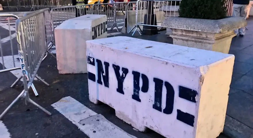 Betonowe bariery NYPD na ulicy. Czy w USA jest bezpiecznie? Widać ludzi i ruch uliczny w tle.