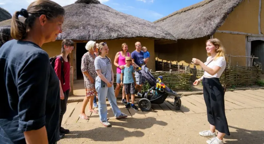 Familienurlaub in Haithabu: Eine Gruppe erkundet historische Häuser mit Strohdächern, während eine junge Frau eine Führung gibt.