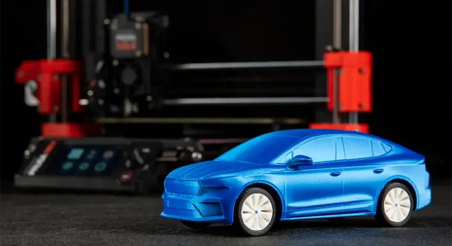 Niebieski model samochodu, gotowy do drukowania, stoi przed drukarką 3D.