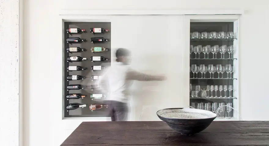 Una nicchia nel muro portante ospita una cantinetta per vini e una vetrina di bicchieri. Una figura sfocata si muove tra i due spazi.
