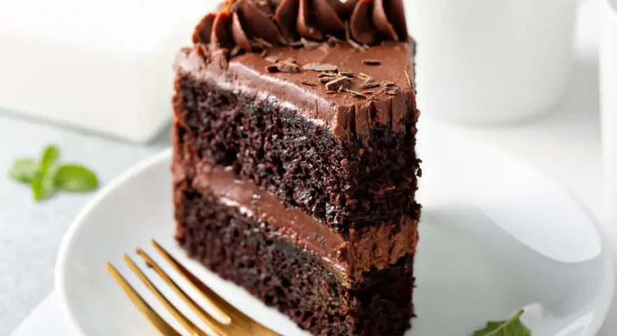 Rebanada de tarta de chocolate con glaseado cremoso y virutas de chocolate. Un tenedor dorado espera pacientemente para probar este delicioso postre.