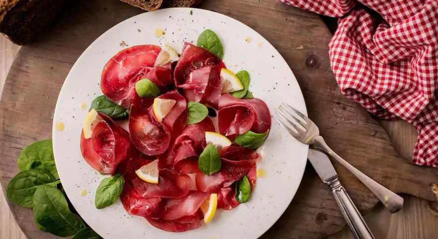 Bresaola affettata finemente, guarnita con spinacini freschi e spicchi di limone. Un'idea semplice su come condire la bresaola per un antipasto leggero.
