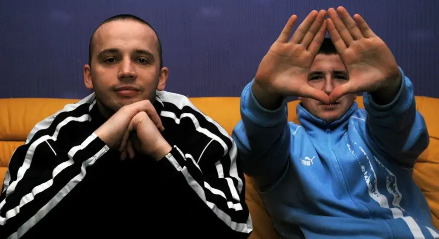 Dwóch mężczyzn w dresach, jeden z rękami złożonymi na brodzie, drugi tworzy rękami kształt serca. Klimat hip hop i rap.