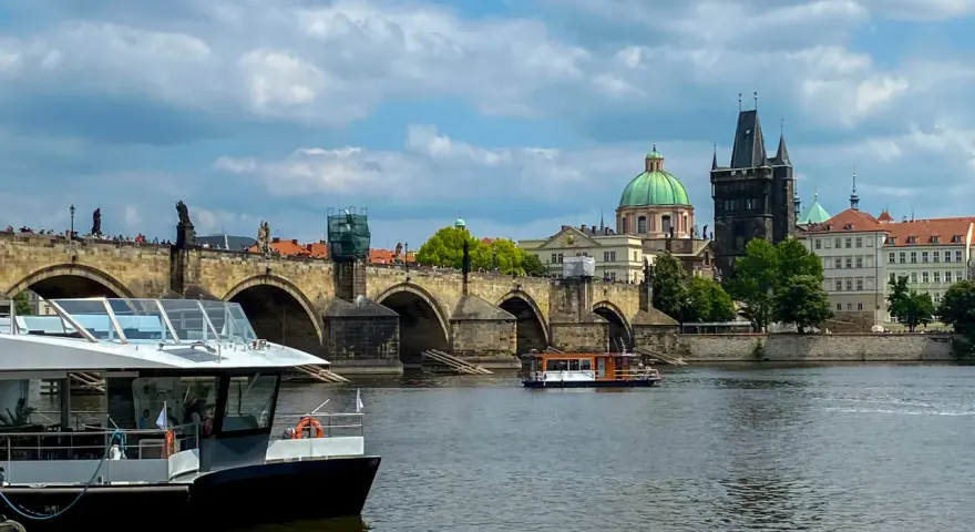 Widok na rzekę w Pradze z łodzią wycieczkową na pierwszym planie i Mostem Karola w tle.