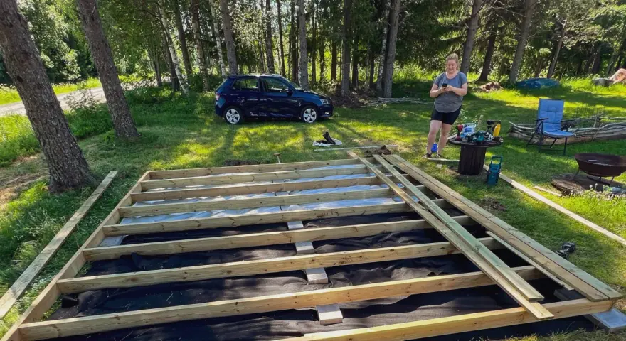 En kvinna förbereder sig för att skruva trall på en nybyggd altan i en lummig trädgård.