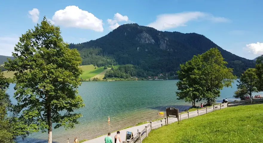 Ein sonniger Tag am bayrischzell see. Menschen spazieren am Ufer entlang, während im Hintergrund ein bewaldeter Berg aufragt.