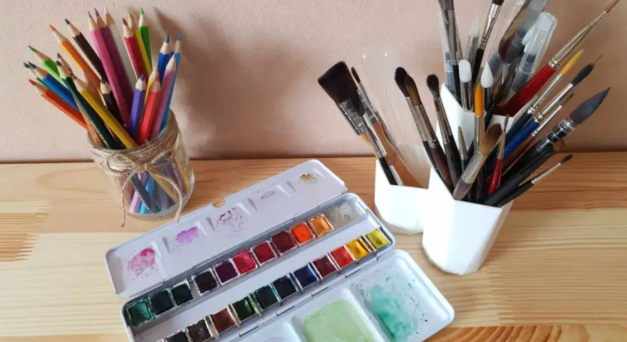 Une palette d'aquarelle ouverte, des crayons de couleur dans un bocal et plusieurs pinceaux indispensables aquarelle dans des pots blancs.
