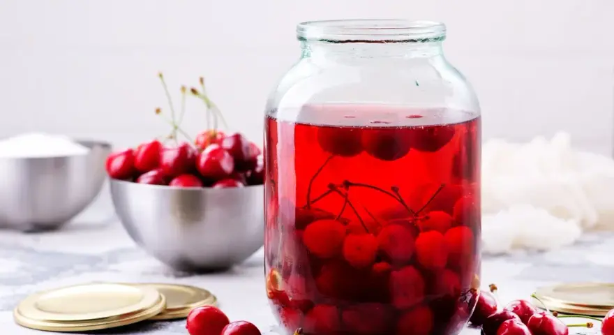 Duży słoik wypełniony rubinowym kompotem z wiśni, obok miska świeżych owoców i cukru.