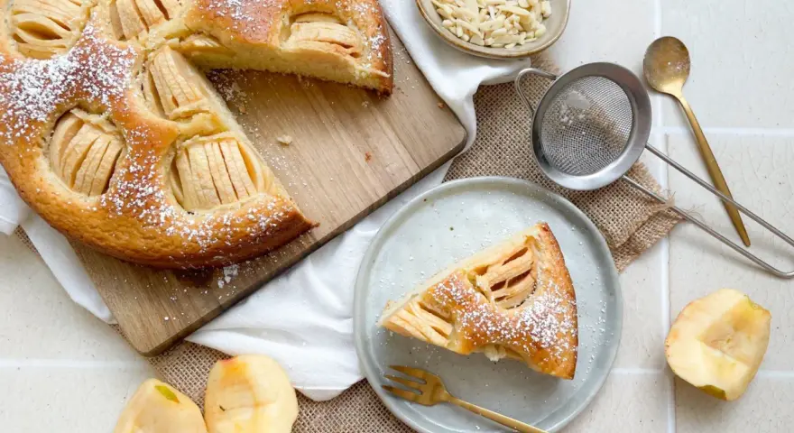 Ein köstlicher veganer Apfelkuchen, bestäubt mit Puderzucker, wird auf einem Holzbrett präsentiert. Daneben liegen frische Apfelstücke und eine Schale mit Mandelblättchen.