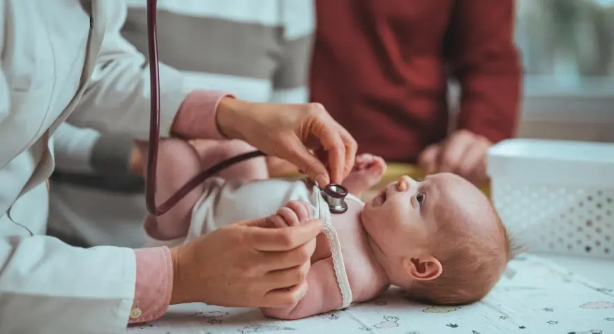 Pielęgniarka środowiskowa po porodzie osłuchuje noworodka. Rodzice obserwują badanie.