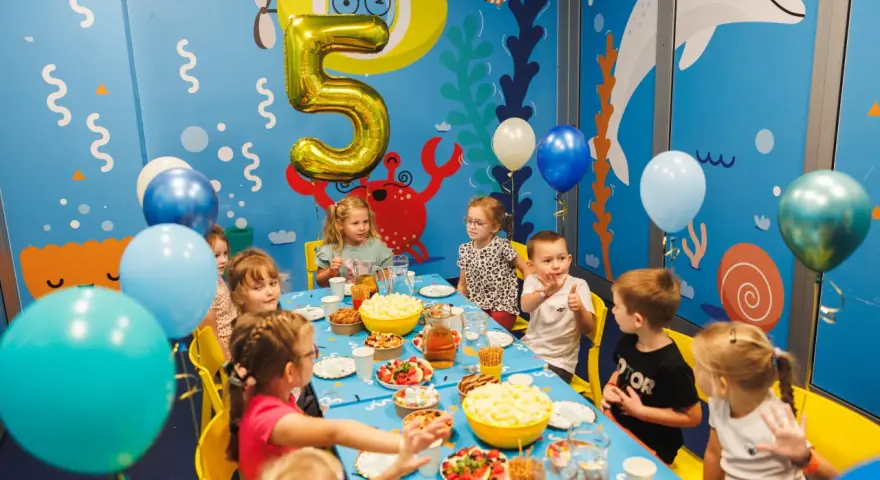 Dzieci świętują 5 urodziny przy stole pełnym przekąsek. Złoty balon w kształcie cyfry 5 unosi się nad stołem.