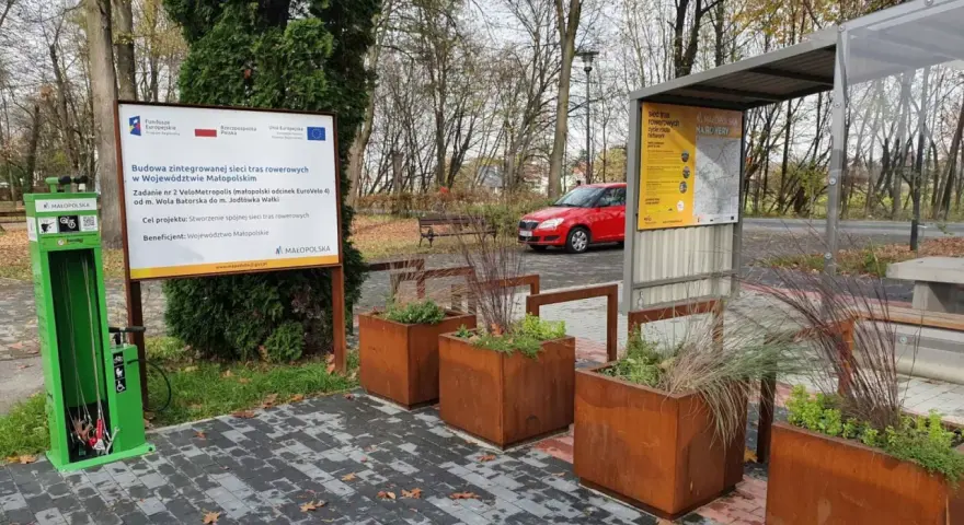 Zielona stacja serwisowa dla rowerów i tablica informacyjna o projekcie VeloMetropolis w Małopolsce. W tle czerwony samochód i jesienne drzewa.