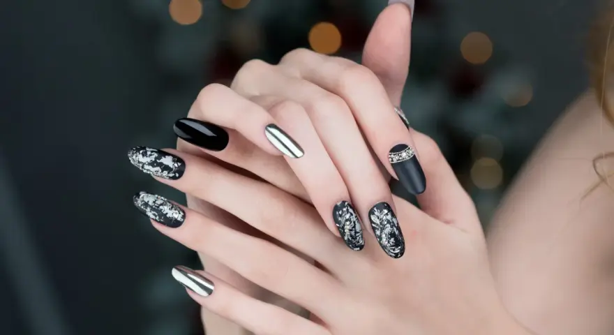 Mani con unghie particolari: nero lucido, argento specchiato e decorazioni floreali in bianco e argento con strass.
