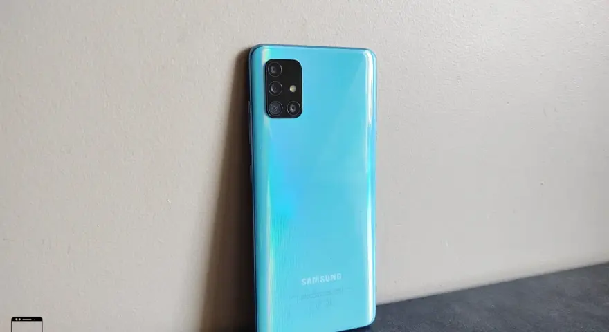 Błękitny Samsung A51 z aparatem w kwadratowej ramce, oparty o jasną ścianę.