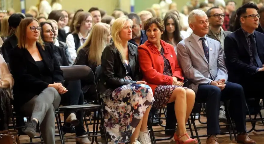 Uczestnicy siedzą na krzesłach, słuchając uważnie. Wśród nich widać osoby, które mogą być na radzie pedagogicznej, w tym kobiety w eleganckich strojach i mężczyzn w garniturach.