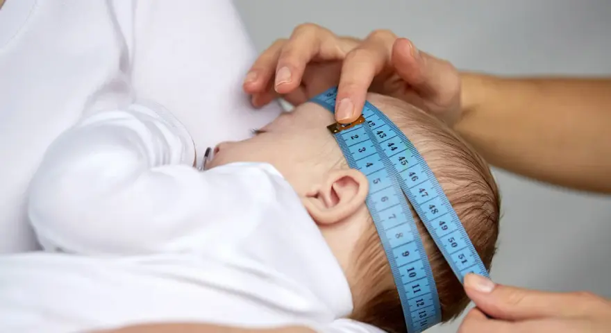 Dłoń mierzy obwód głowy niemowlęcia niebieską miarką krawiecką.