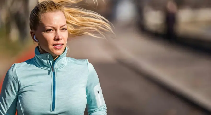 Kvinna med blont hår i hästsvans springer utomhus, lyssnar på musik med hörlurar. Hon är fokuserad på att springa 5 km tid.