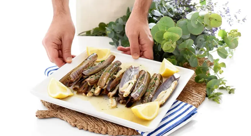 Un plato blanco con navajas a la plancha, limón y perejil. Unas manos sostienen el plato, decorado con eucalipto y flores moradas.