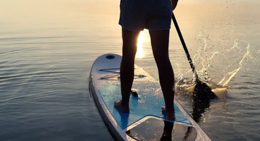 Person lernt SUP auf ruhigem Wasser bei Sonnenuntergang. Das Paddel spritzt Wasser auf.