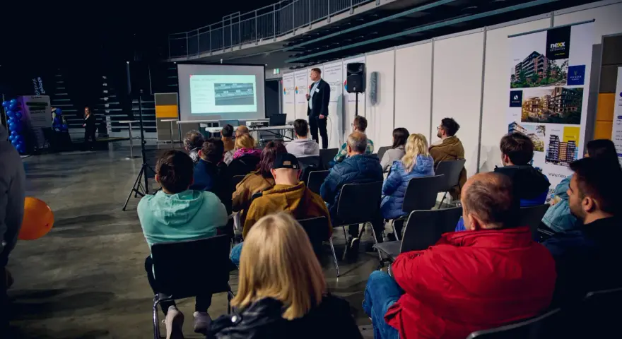 Prezentacja na targach budowlanych. Mężczyzna przemawia do zgromadzonej publiczności, która siedzi na krzesłach.