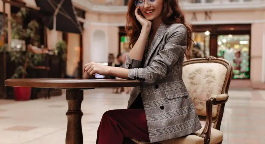 Elegancka kobieta w okularach i marynarce w kratę, prezentująca stylizacje z bordowymi spodniami, siedzi przy stoliku w kawiarni.