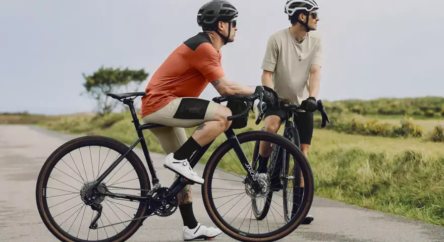 Dwóch rowerzystów na szosie, jeden w pomarańczowej koszulce, drugi w beżowej. To idealny widok na to, co to gravel - wszechstronność i przygoda.