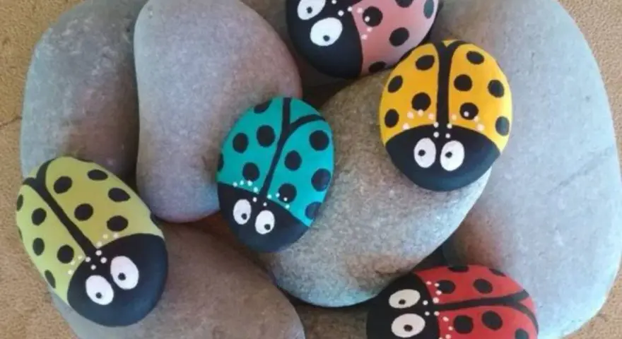 Seis piedras pintadas fáciles con mariquitas de colores: roja, amarilla, verde, rosa y turquesa, descansan sobre piedras grises.