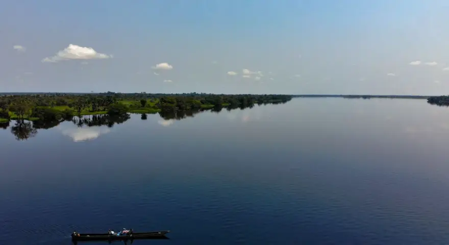 Szeroka, spokojna rzeka Kongo, odbijająca niebo i bujną zieleń brzegów. Na wodzie płynie łódź z kilkoma osobami.