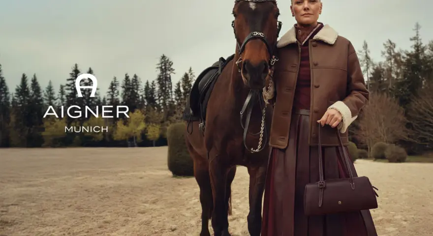 Elegancka kobieta w brązowej kurtce i spódnicy obok konia. Logo AIGNER MUNICH. Aigner co to za marka? Luksusowa moda.