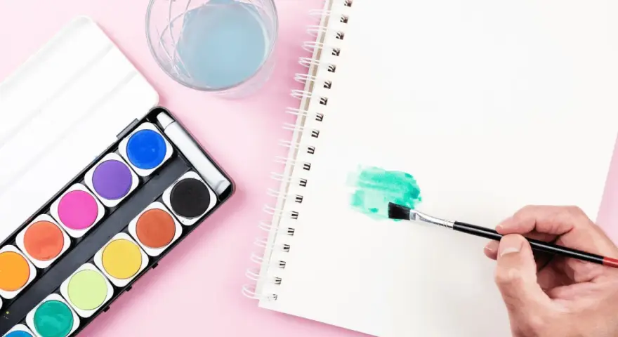 Une main tient un pinceau pour créer une tache verte sur un carnet, à côté d'une palette d'aquarelle et d'un verre d'eau.