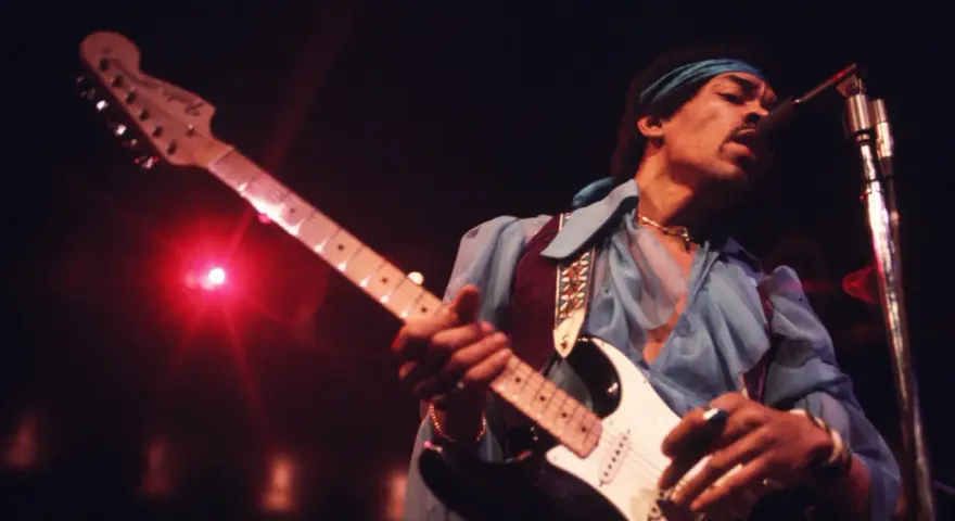 Jimi Hendrix, einer der besten Gitarristen, spielt auf der Bühne seine weiße Fender Stratocaster. Rotes Licht beleuchtet die Szene.