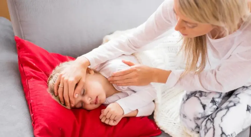 Matka troskliwie opiekuje się dzieckiem, które leży na czerwonej poduszce. Dziecko ma zamknięte oczy, a matka delikatnie dotyka jego czoła, sprawdzając, czy nie ma gorączki. To obrazek pokazujący troskę o dziecko podczas jelitówki.