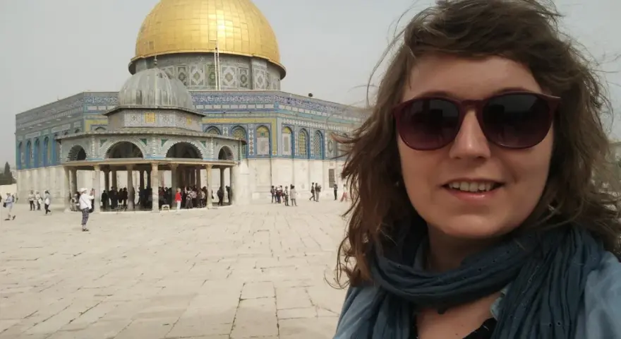 Uśmiechnięta kobieta w okularach przeciwsłonecznych robi selfie na tle Kopuły na Skale. Wokół widać turystów, co sugeruje, że w Jerozolimie jest bezpiecznie.