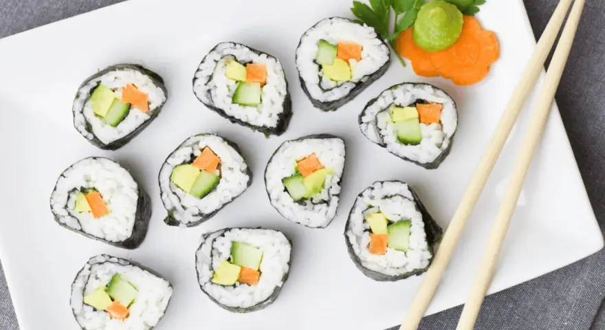 Talerz z rolkami sushi z awokado, ogórkiem i marchewką. Obok pałeczki i dekoracja z marchewki z wasabi. Idealna ilość octu do sushi podkreśla smak.