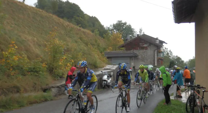 Ciclisti in gara su una strada bagnata, con una **forcella alta** che indica la salita. Il paesaggio è autunnale, con alberi e un pendio erboso.