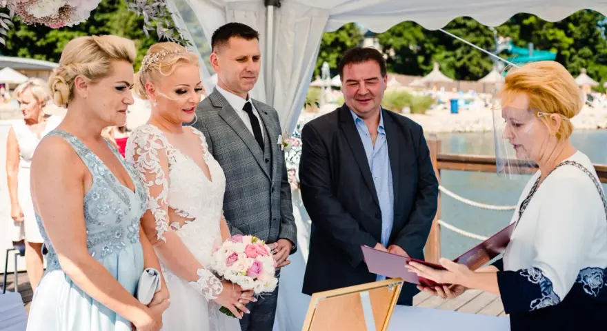 Ceremonia ślubna nad jeziorem, panna młoda w białej sukni, pan młody w garniturze, urzędniczka w przyłbicy.