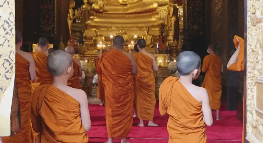 A group of Buddhist monks, another word for ascetics, walk towards a large golden Buddha statue in a temple.