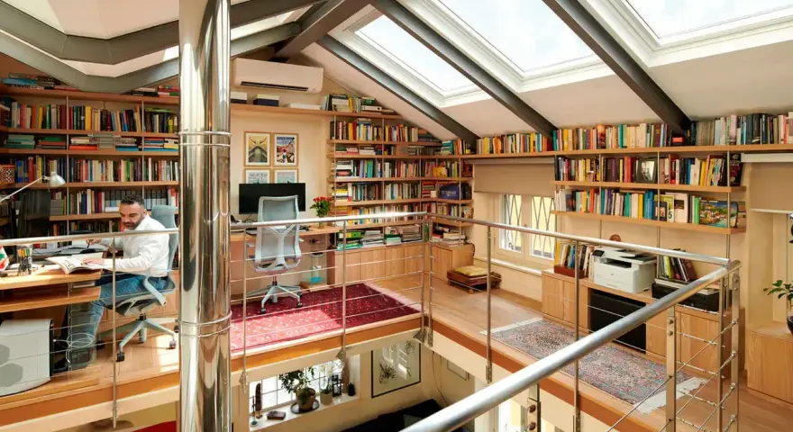 Un uomo lavora in una luminosa libreria mansarda, circondato da scaffali pieni di libri e illuminato da lucernari.