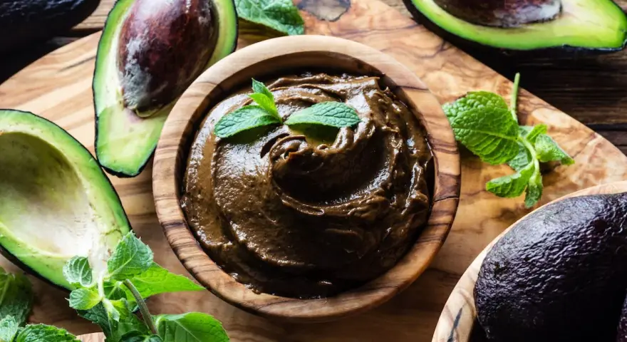 Crema di avocado vellutata in ciotola di legno, circondata da avocado freschi e foglie di menta.