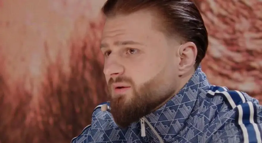 Amadeusz Ferrari w niebieskiej bluzie z białymi paskami, z charakterystyczną fryzurą i brodą, siedzi przed ekranem.