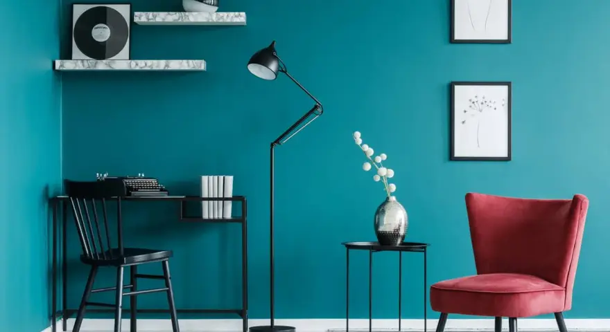 Bureau noir avec machine à écrire, lampe sur pied, fauteuil rouge et étagères sur un mur bleu ou vert canard.