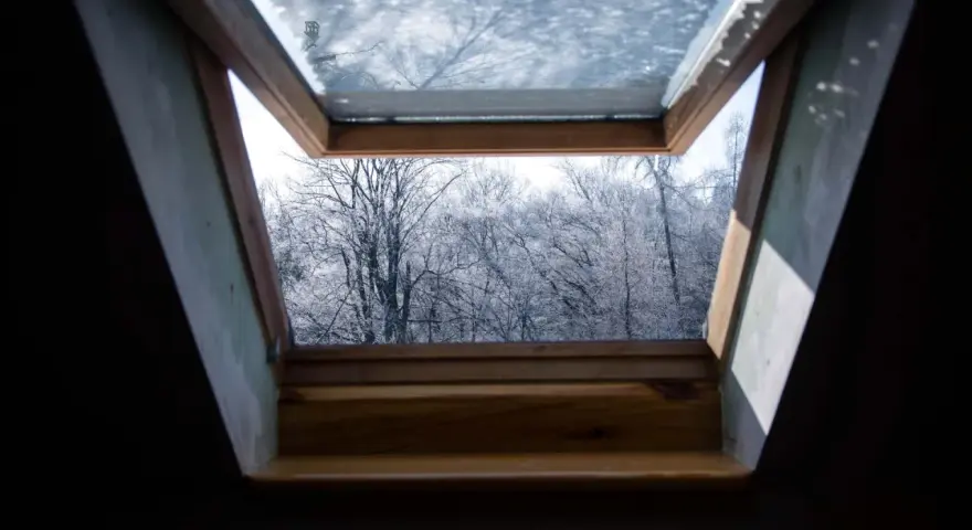 Ein geöffnetes Kellerfenster im Winter gibt den Blick frei auf kahle, schneebedeckte Bäume.